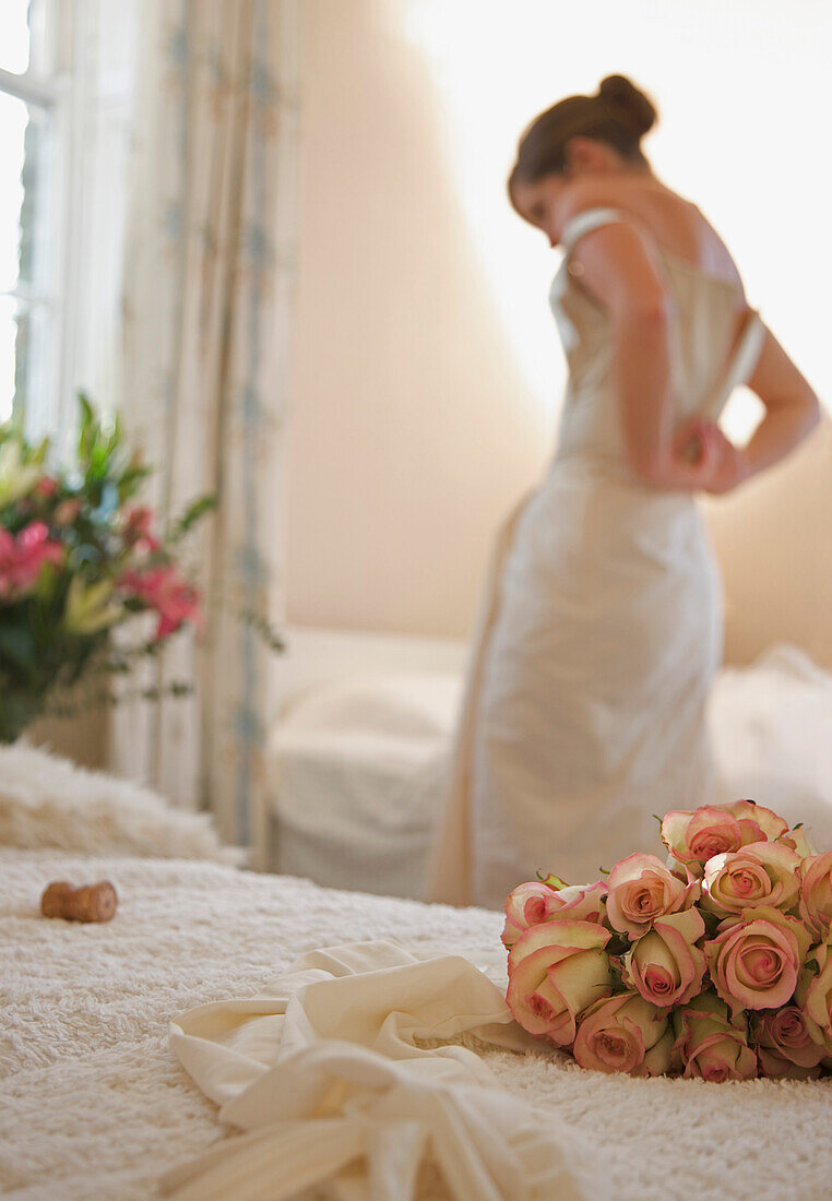 Nahaufnahme eines Rosenstraußes und von Brauthandschuhen, die auf einem Bett liegen, während sich die Braut entkleidet