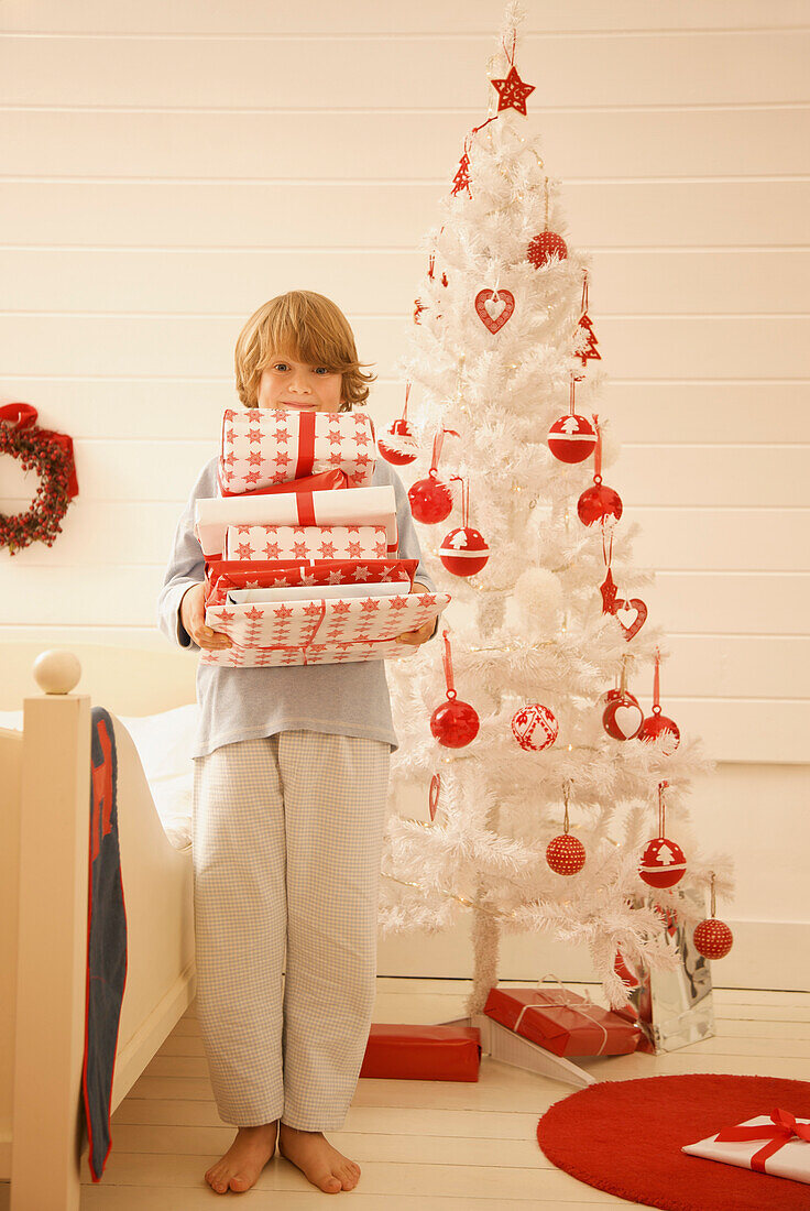 Junge steht an einem Weihnachtsbaum und hält einen Stapel Geschenke in der Hand