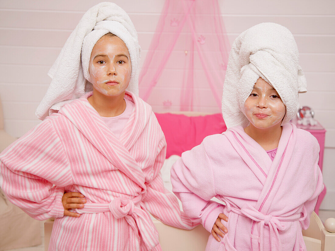Zwei Mädchen mit Gesichtsmasken, Handtuchturbane und Morgenmäntel, die mit den Händen in den Hüften posieren