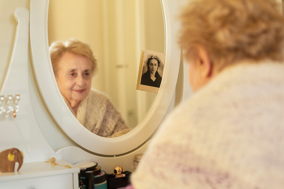 Lächelnde ältere Frau betrachtet ihr Spiegelbild neben einem alten Foto