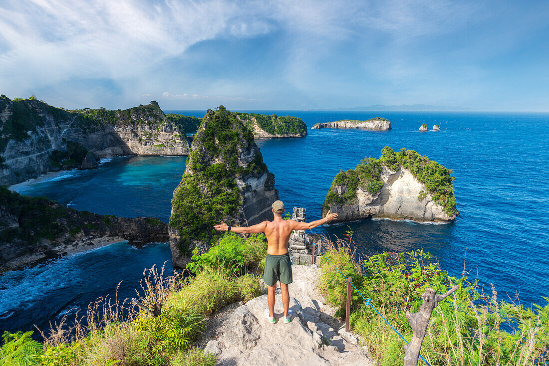 Rückansicht eines jungen Mannes mit ausgestreckten Armen, der auf einer Klippe mit Blick auf den Ozean steht, Diamond Beach, Nusa Penida, Klungkung regency, Bali, Indonesien, Südostasien, Asien