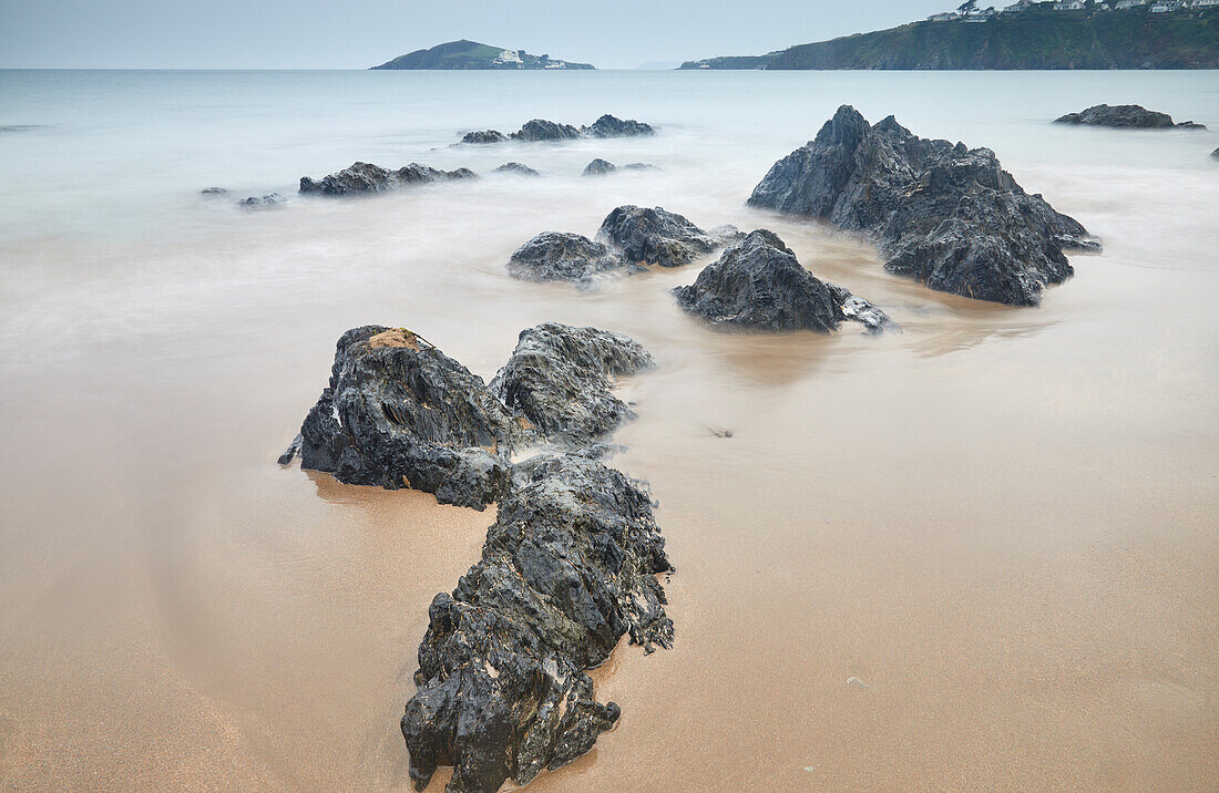 In Sand eingegrabene Felsbrocken an der Küste in der Abenddämmerung, Blick vom Strand von Bantham in Richtung Burgh Island, an der Südküste von Devon, England, Vereinigtes Königreich, Europa