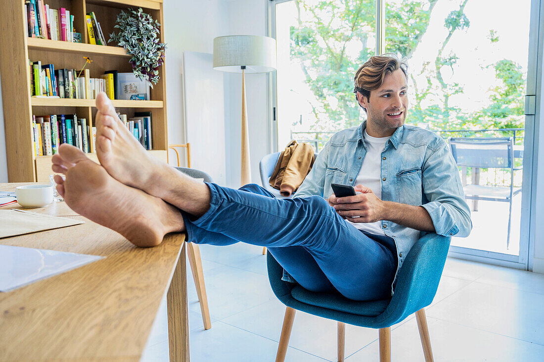 Young adult man resting feet while working with smart phone at home