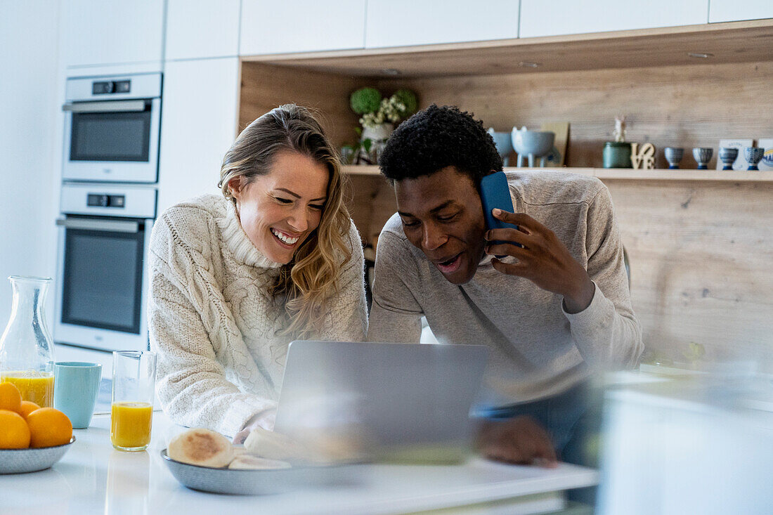 Cheerful couple leaning on kitchen counter using laptop