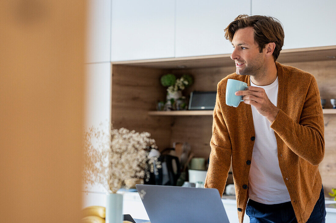 Nachdenklicher junger Mann trinkt eine Tasse Kaffee, während er in der Küche steht