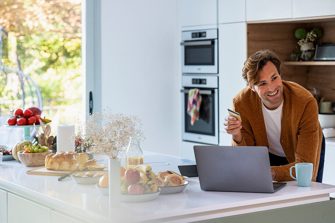 Erwachsener Mann beim Online-Shopping am Laptop in der Küche