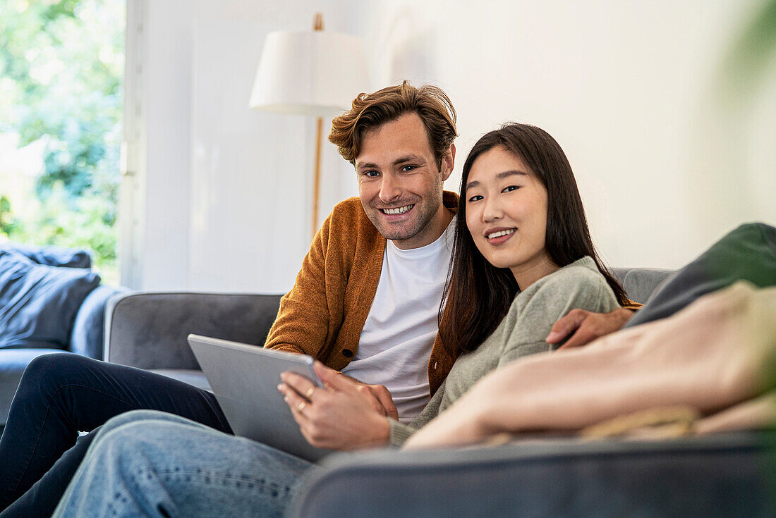 Erwachsenes Paar schaut in die Kamera, während es auf dem Sofa sitzt und ein digitales Tablet benutzt