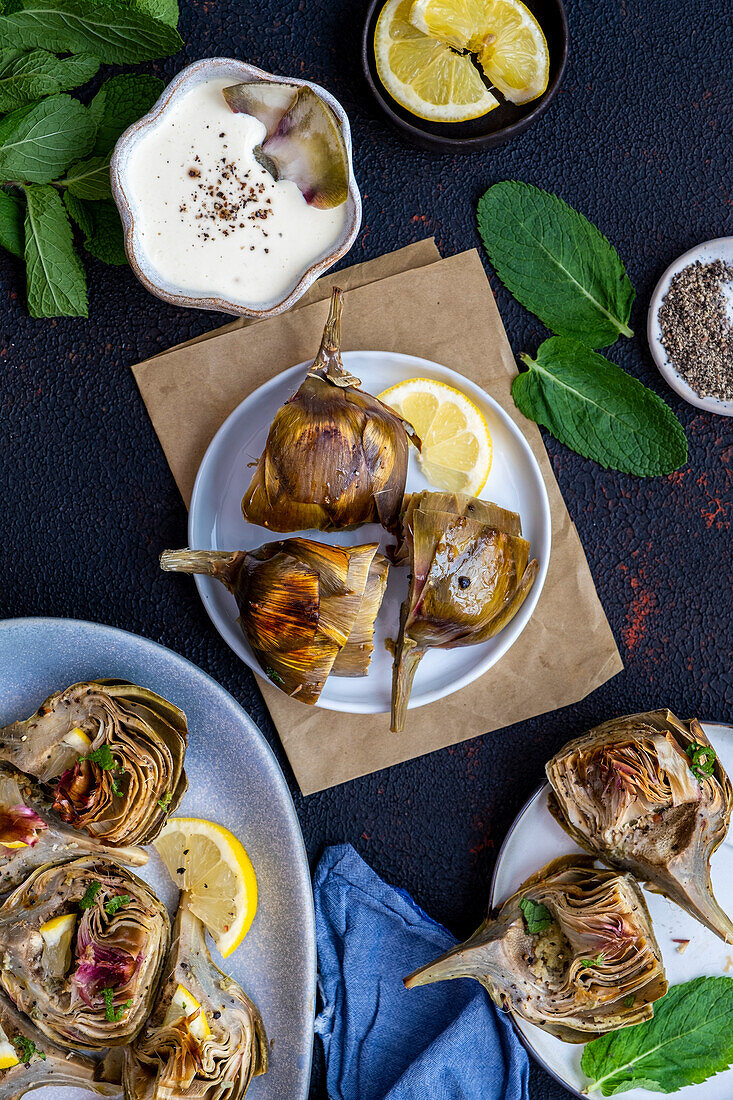 Roasted artichokes served on a white plate, accompanied by a bowl of dipping sauce, mint leaves and lemon slices.
