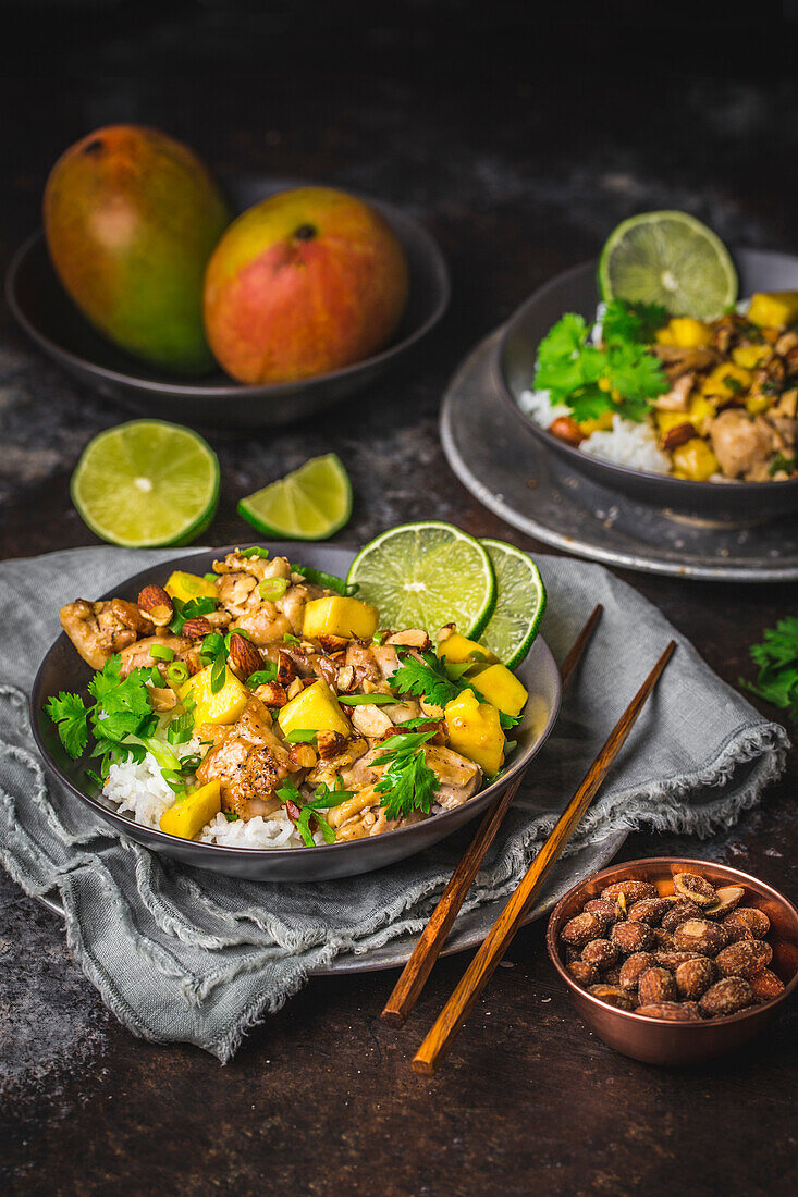 Gebratenes Hähnchen und Mango mit Limettenscheiben und Koriander, in dunklen Schalen mit Reis und Stäbchen, Mangos im Hintergrund