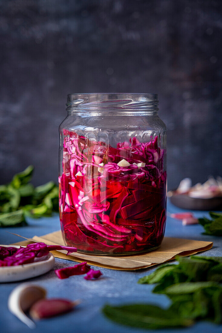 Red cabbage pickles in a jar on a dark backdrop and garlic cloves, herbs around it.