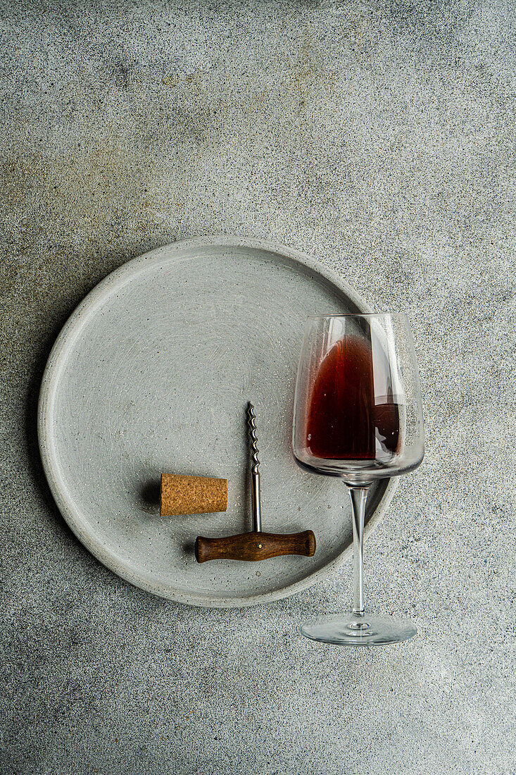 Hoher Blickwinkel auf ein Glas trockenen Rotweins, der auf einem Teller mit Korkenzieher und Korken auf einem Betonhintergrund liegt