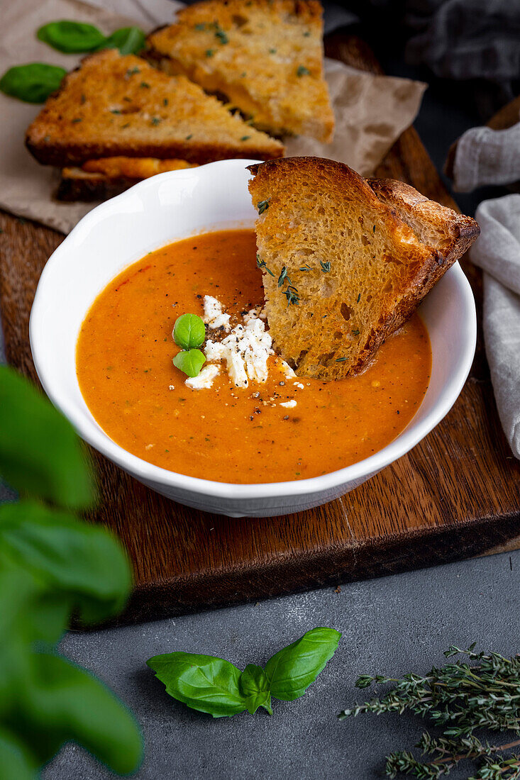 Roasted tomato soup with feta cheese and basil leaves in a white bowl, grilled cheese sandwich dipped in it and more grilled cheese sandwiches behind it