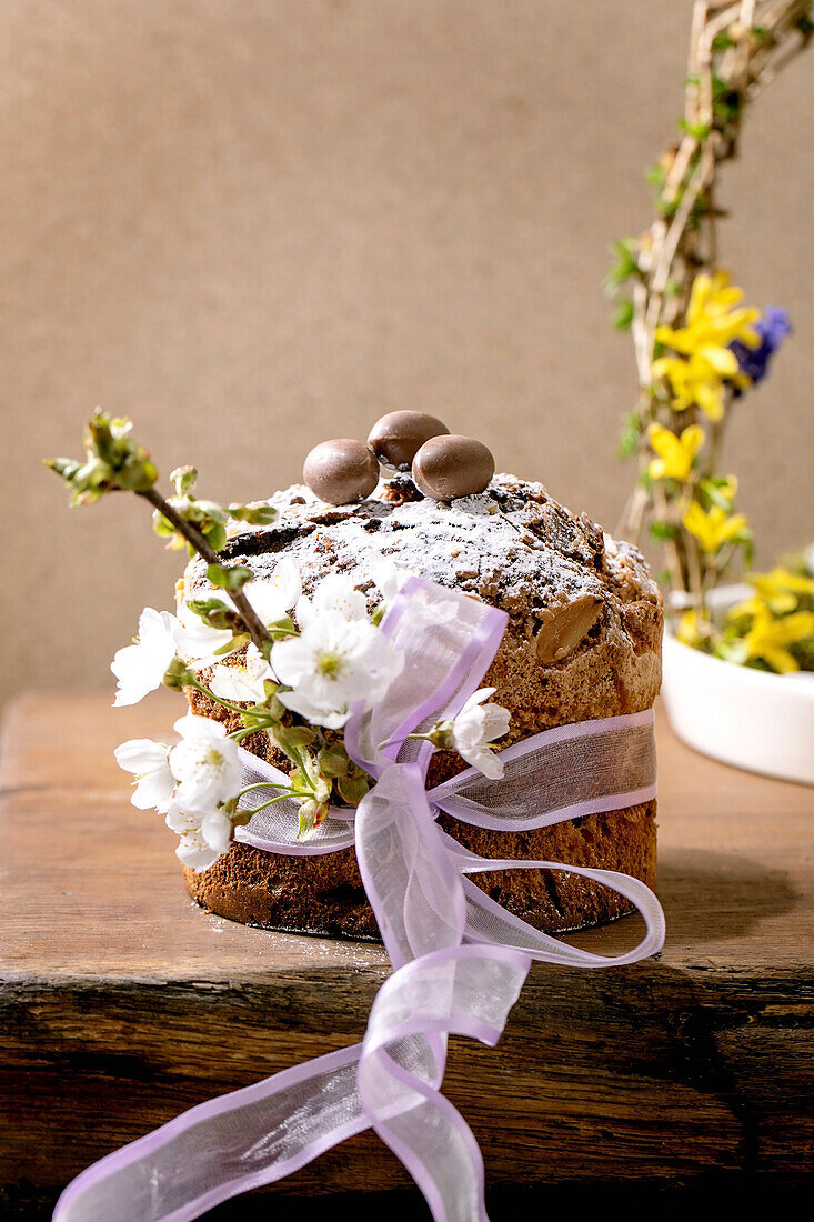 Homemade Italian traditional Easter panettone cake, decorated by chocolate eggs, pink ribbon and blossoming cherry tree flowers standing on wooden table. Traditional Easter European bake. Copy space