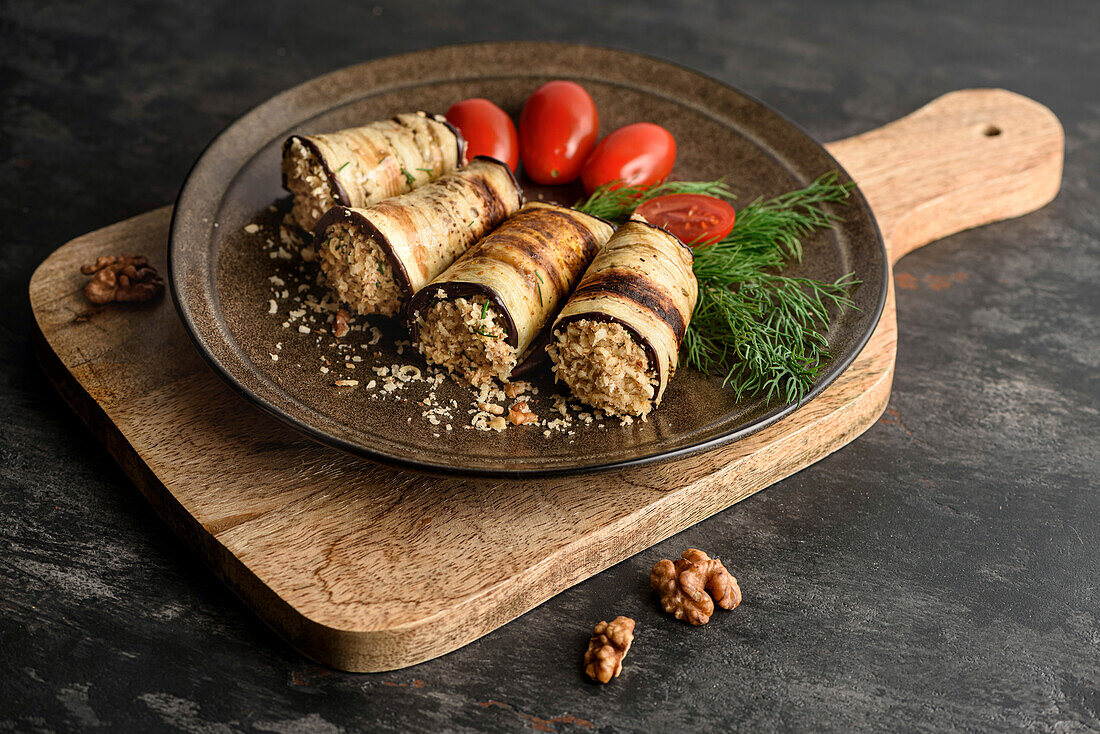 Gegrillte Auberginenröllchen mit Walnussfüllung, Tomaten und frischem grünen Dill. Der Teller steht auf dem Schneidebrett