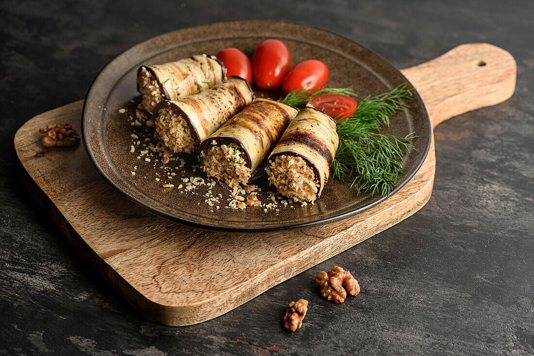 Grilled aubergine rolls with walnut filling, tomatoes and fresh green dill. The plate is on the chopping board