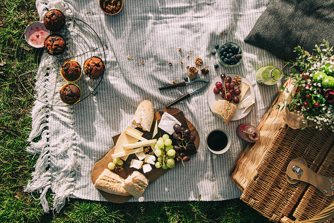 Flatlay picnic scene with a basket and flowers, juice, grapes, cheese and baguette