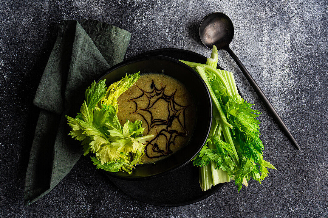 Top view of healthy celery cream soup in bowl with celery sticks served on black plate with black spoon and napkin against blurred dark background