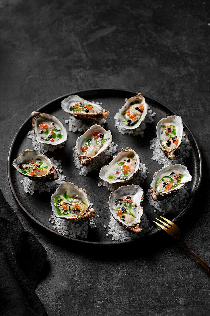 Freshly shucked oysters with buttermilk dressing, salmon roe, balsamic pearls and herb oil on rock salt on a black plate