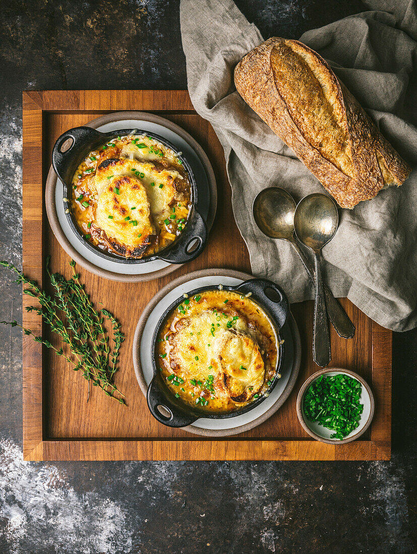 Two bowl of Onion Soup in cast iron bowls topped with melted cheese, on wood tray with bread, spoons and chives