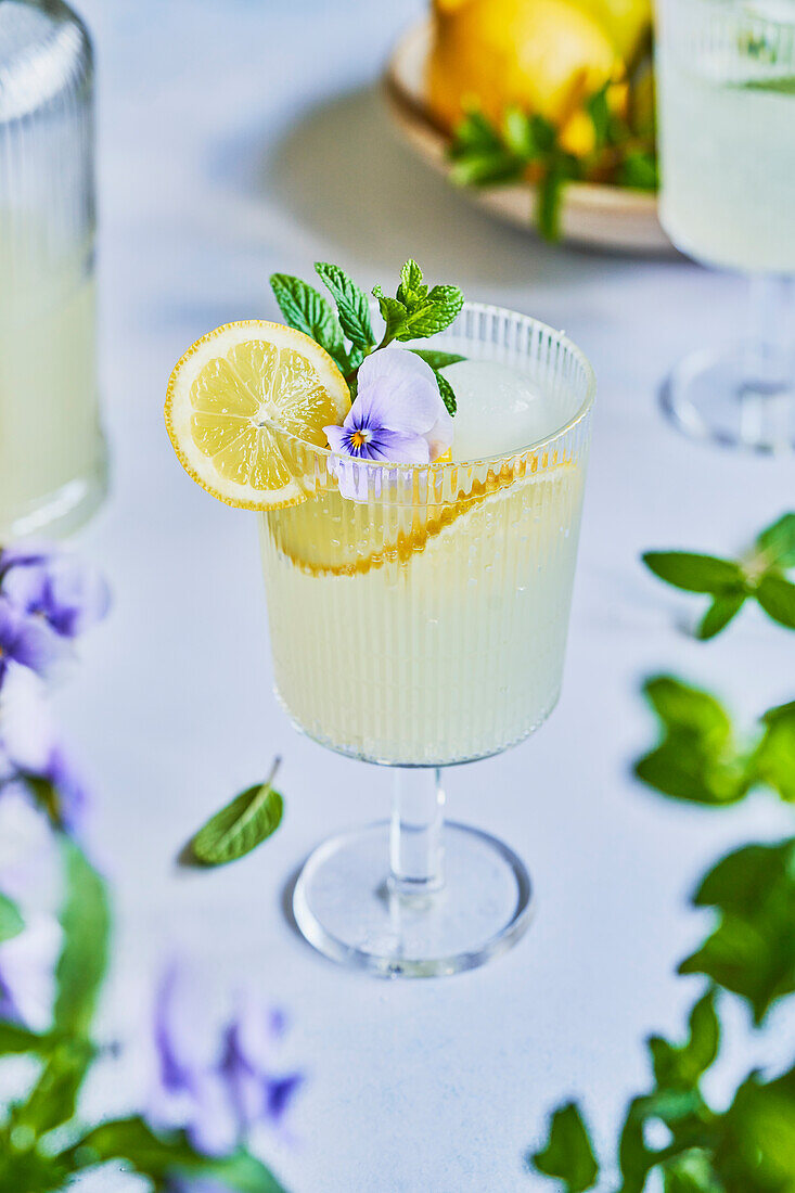 Lemon and mint lemonade mocktail on a light blue background with lemon, mint and purple flower garnish