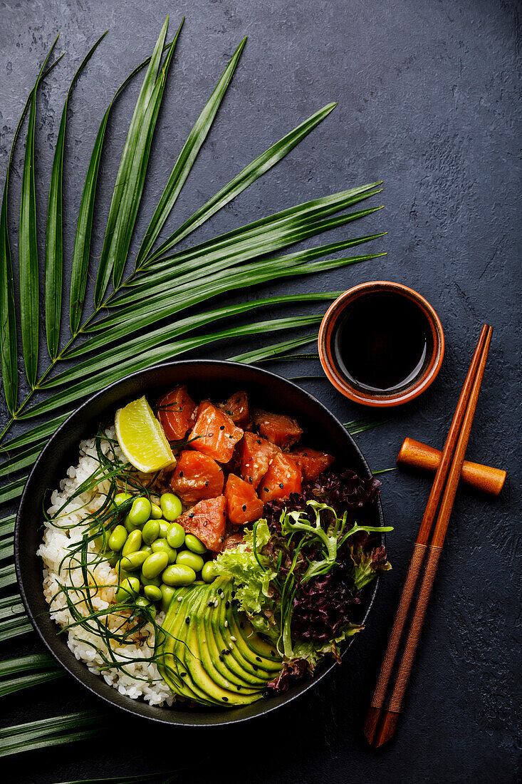 Salmon Poke Bowl Raw fish salad Asian trend dish with soya beans, edamame, rice, avocado and salad in a bowl on a tropical leaf and dark background