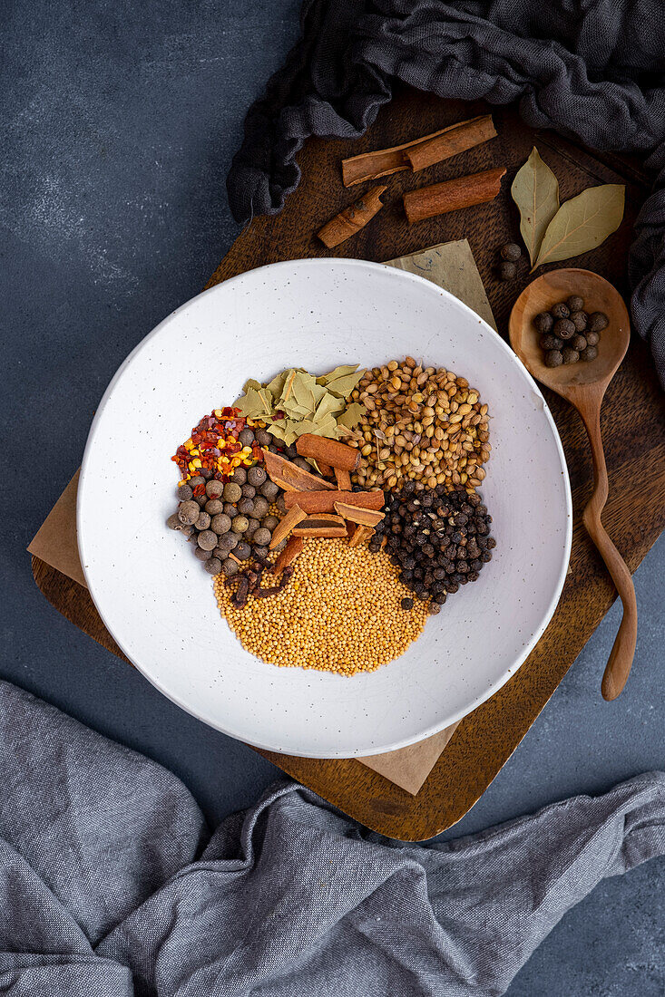 Mustard seeds, coriander seeds, cloves, cinnamon sticks, bay leaves and red pepper flakes, mixed in a white ceramic bowl on a wooden board
