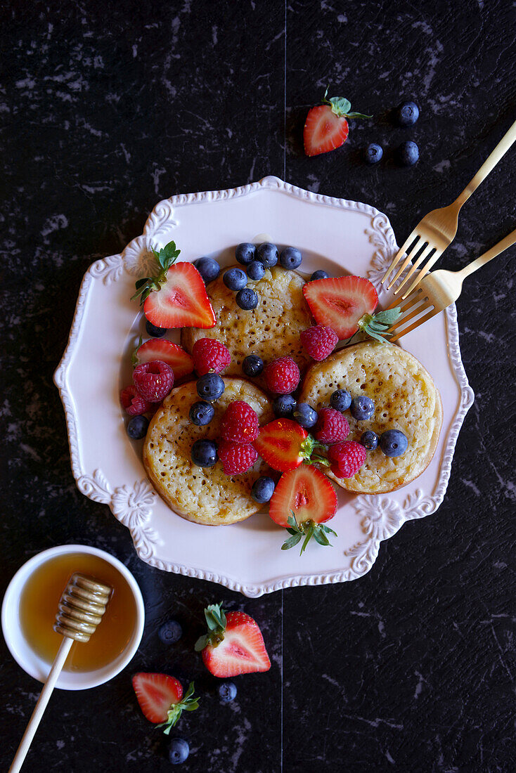 English style crumpets served with berries and honey on black marble background, flatlay close up.