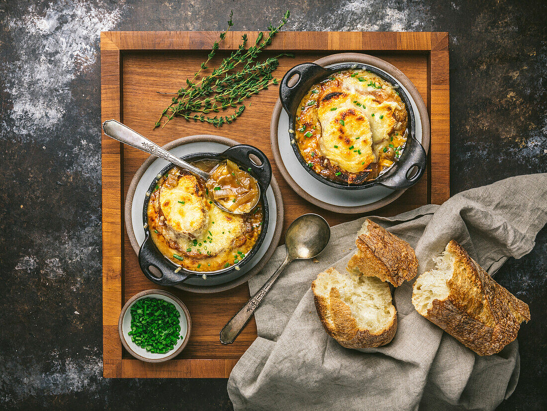 Two bowls of onion soup in cast-iron bowls with melted cheese, spoon in bowl, on wooden tray with bread and chives