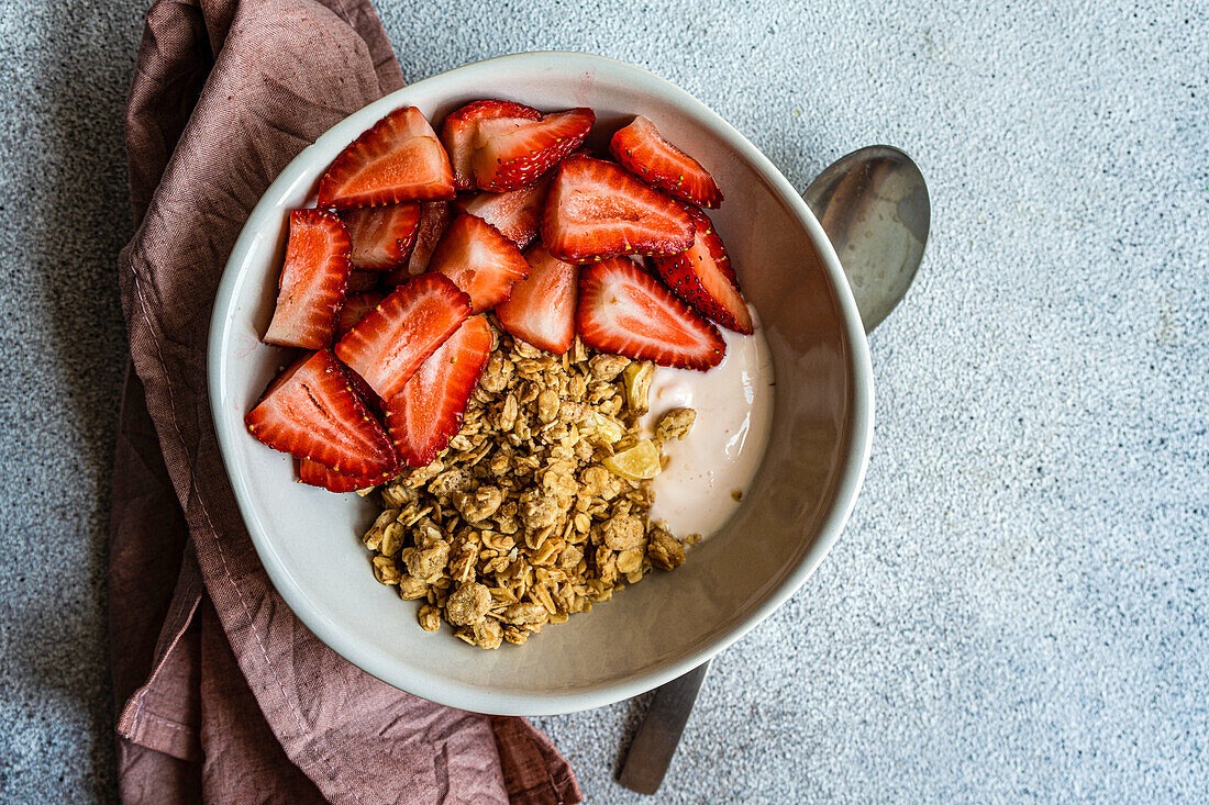 Bowl with muesli, yoghurt and ripe organic strawberries on a grey background between spoon and pink napkin seen from above