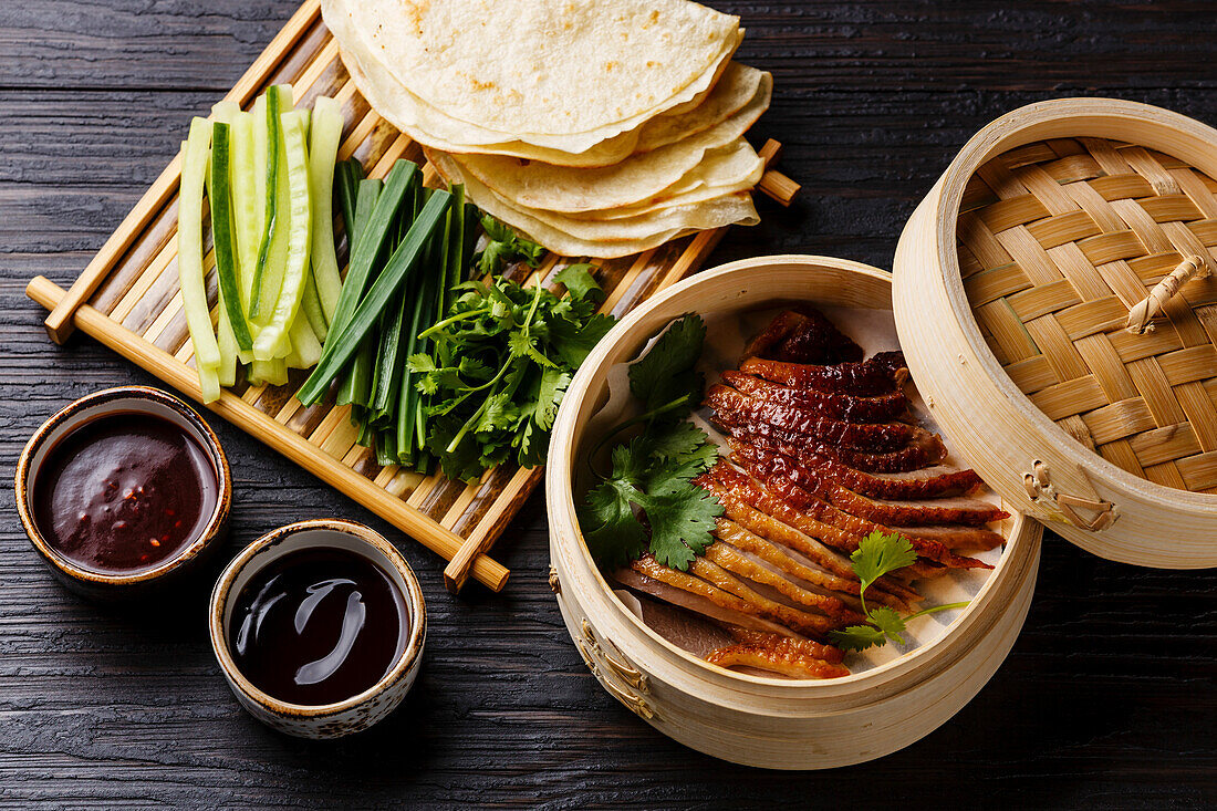 Peking Duck in bamboo steamer served with fresh cucumber, green onions, cilantro and roasted wheaten chinese pancakes with sauce Hoysin on black burned wooden background