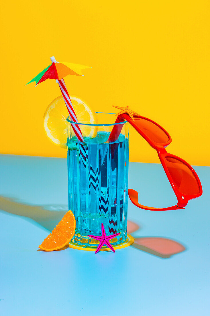 Komposition aus Glas mit blauer Flüssigkeit, Zitronenscheibe und Strohhalm mit stilvoller roter Brille auf hellblauem und gelbem Hintergrund im Studio