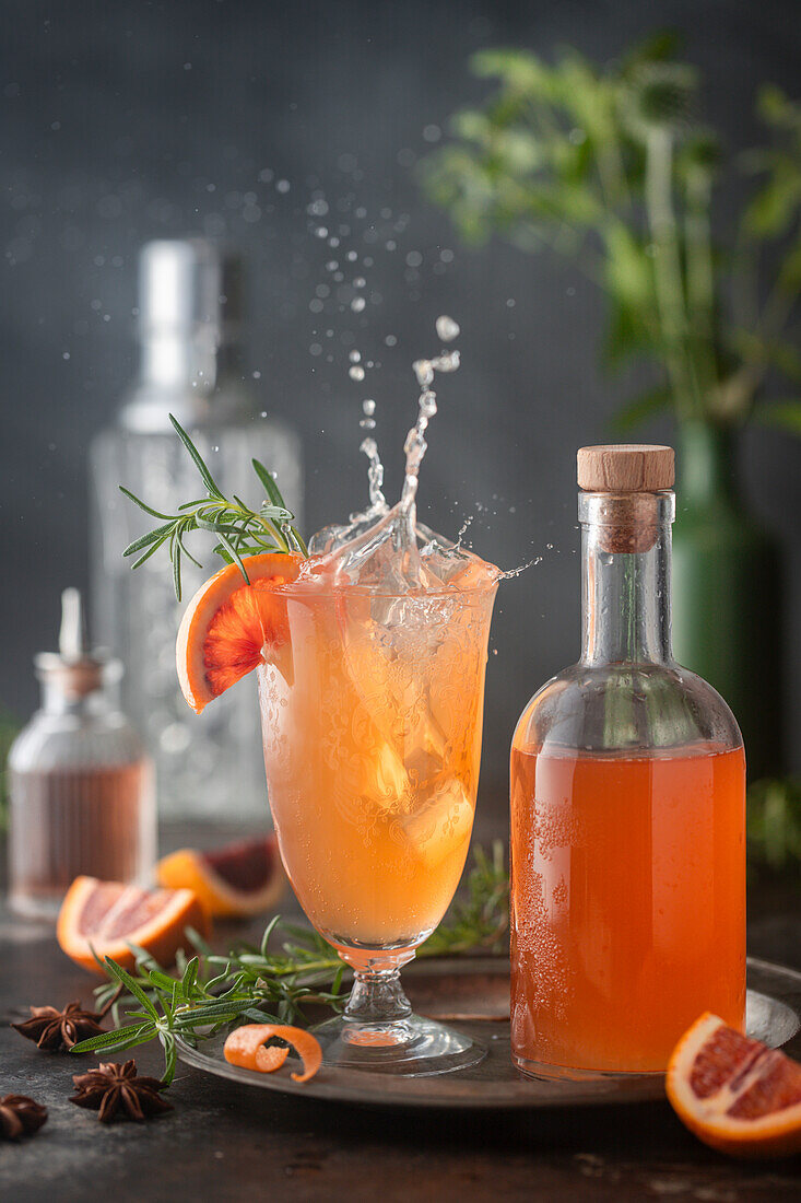 Action-Splash-Shot mit Eis, das in einen Blutorangen-Cocktail im Vintage-Glas fällt, mit Sirupflasche und Zitrus- und Rosmaringarnitur