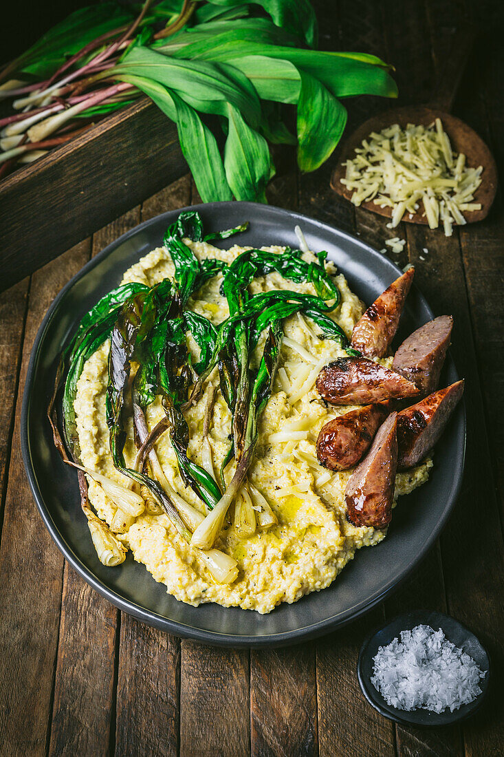 Polenta, grilled ramps and sliced sausage on a platter with fresh ramps and grated cheese in background
