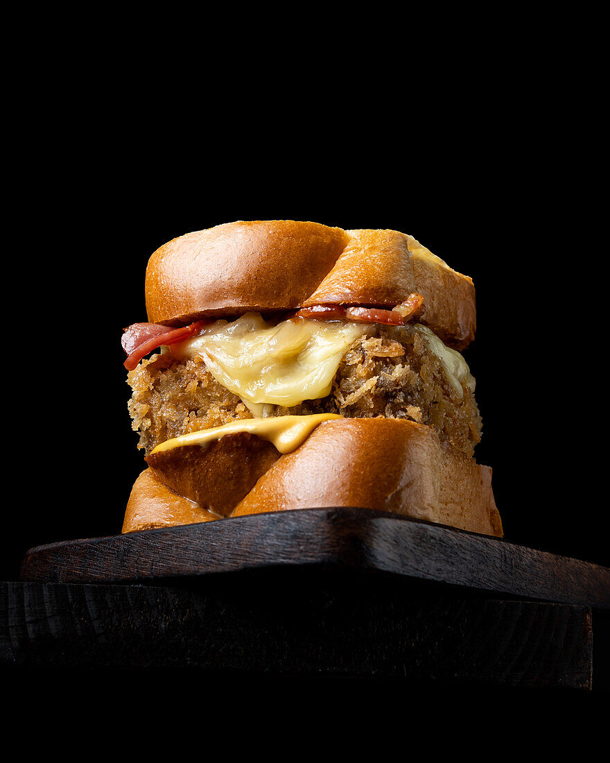 From below appetizing burger with fresh bread cheese and bacon served on a wooden board on a black background