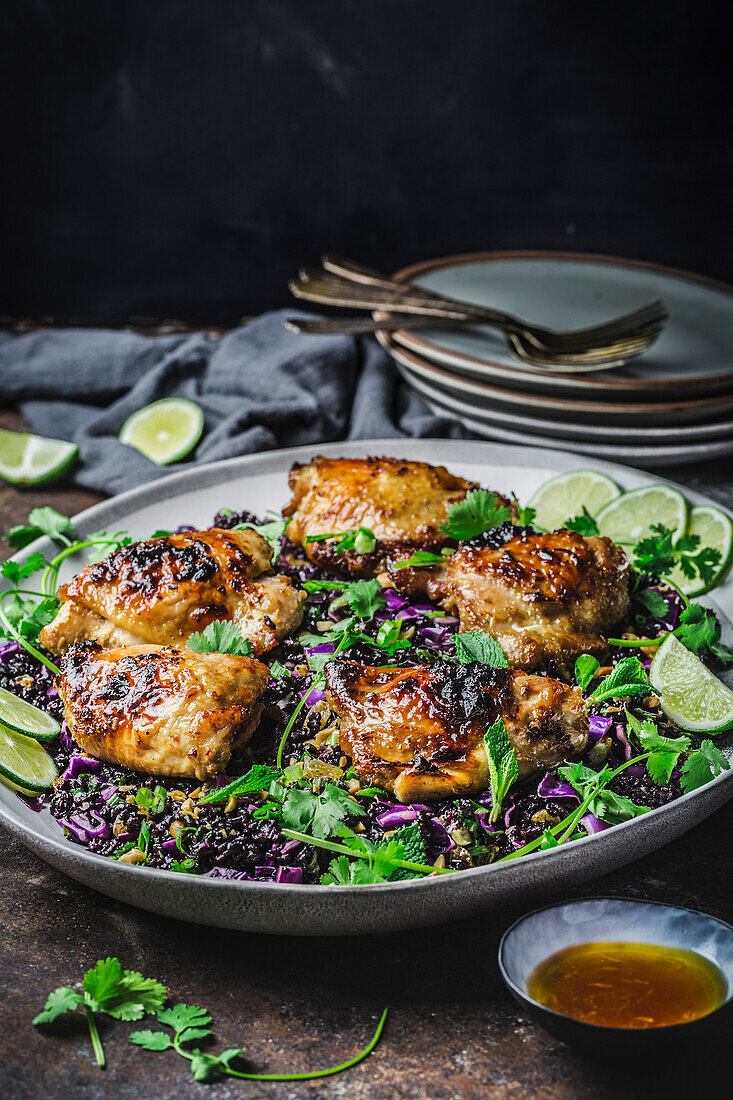 Glazed fried chicken pieces on black rice, red cabbage salad on an oval platter and plates in the background