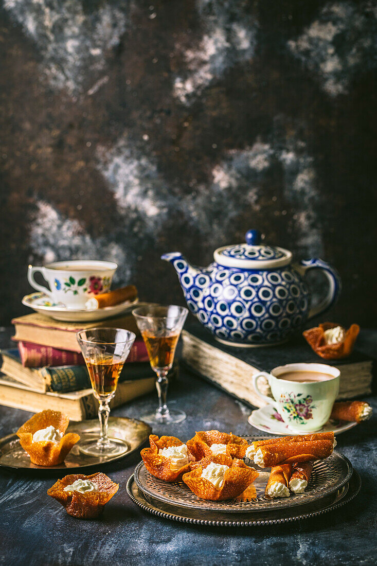 Silbertablett mit einer Reihe von Brandy-Snap-Keksen, gefüllt mit Schlagsahne, und Sherry in alten Gläsern, Teetassen und blau-weißer Teekanne, arrangiert auf antiken Büchern