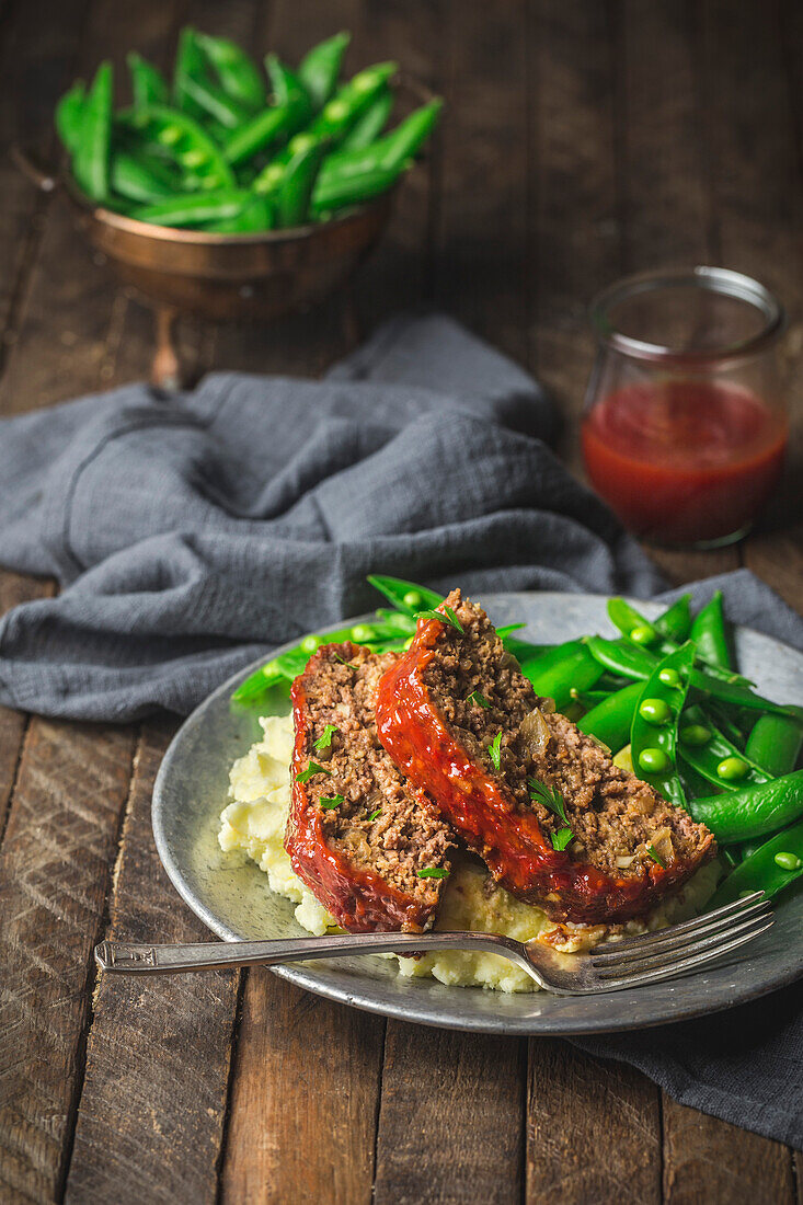 Meatloaf slices on plate with mashed potato, fresh snap peas and sauce