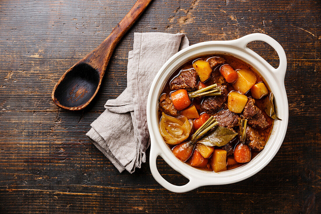 Beef meat stewed with potatoes, carrots and spices in ceramic pot on wooden background