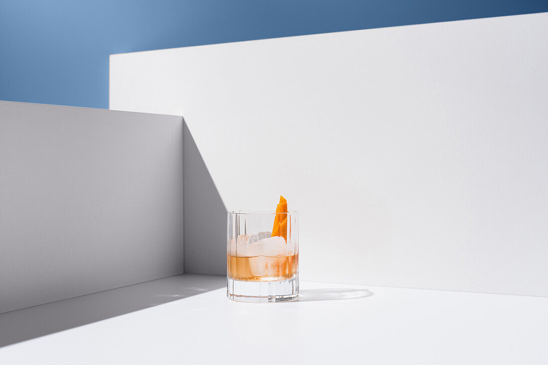 Crystal clear glass filled with fresh old-fashioned cocktail garnished with oranges and ice cubes on a white surface between white walls against a grey background