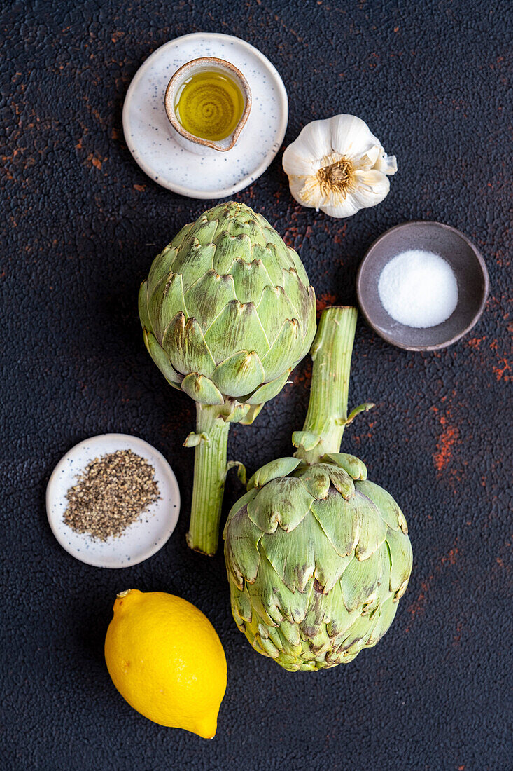 Artischocken, Olivenöl, Knoblauch, Salz, Pfeffer und Zitrone auf dunklem Hintergrund, von oben fotografiert