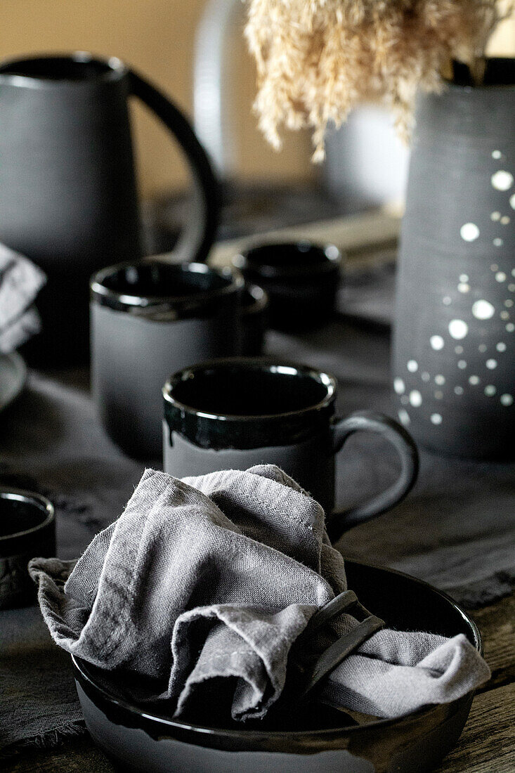 Rustikaler Tisch mit leerem handgefertigtem Keramikgeschirr, schwarzen Schüsseln, Tellern, Tassen, Krug und Vase auf Leinentischdecke. Blck Keramik Rosen Blumen Dekor. Tageslicht Esszimmer