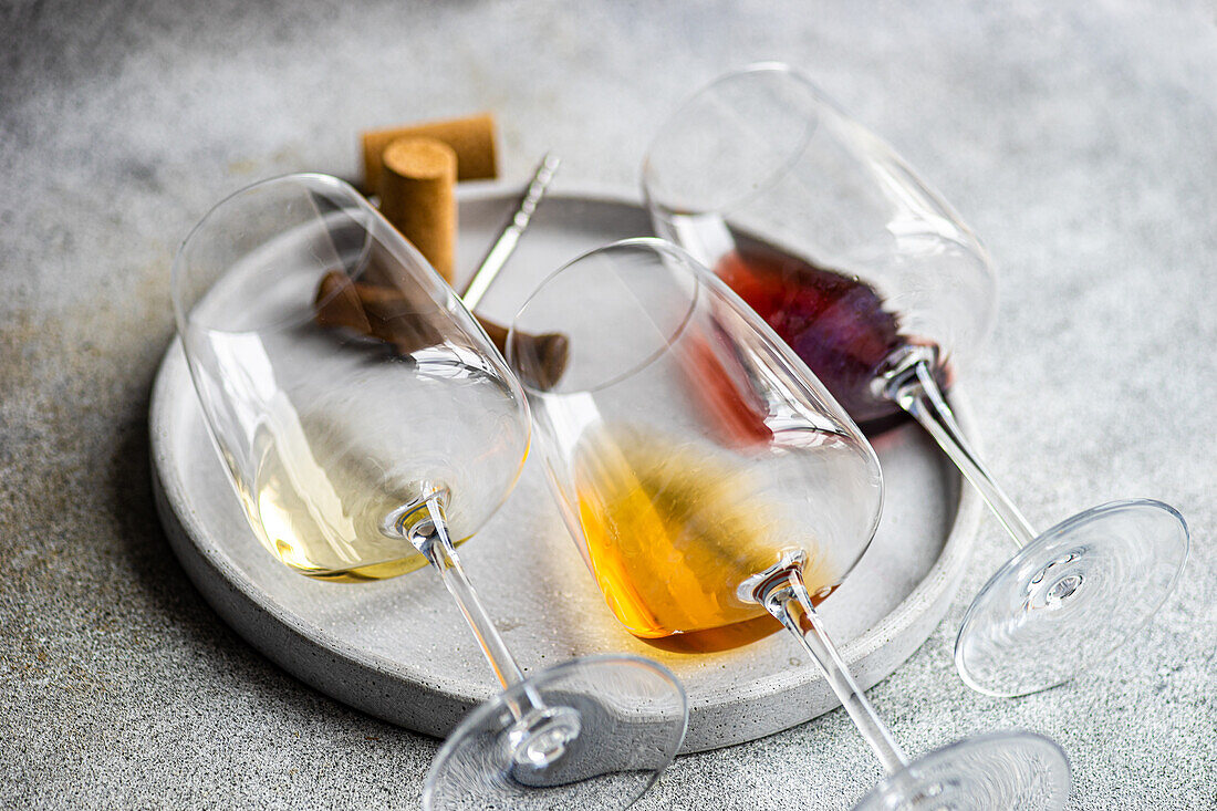 Front view of three varieties of Georgian dry wine glasses (white, amber and red) lying on a plate with a corkscrew and a cork on concrete grey table