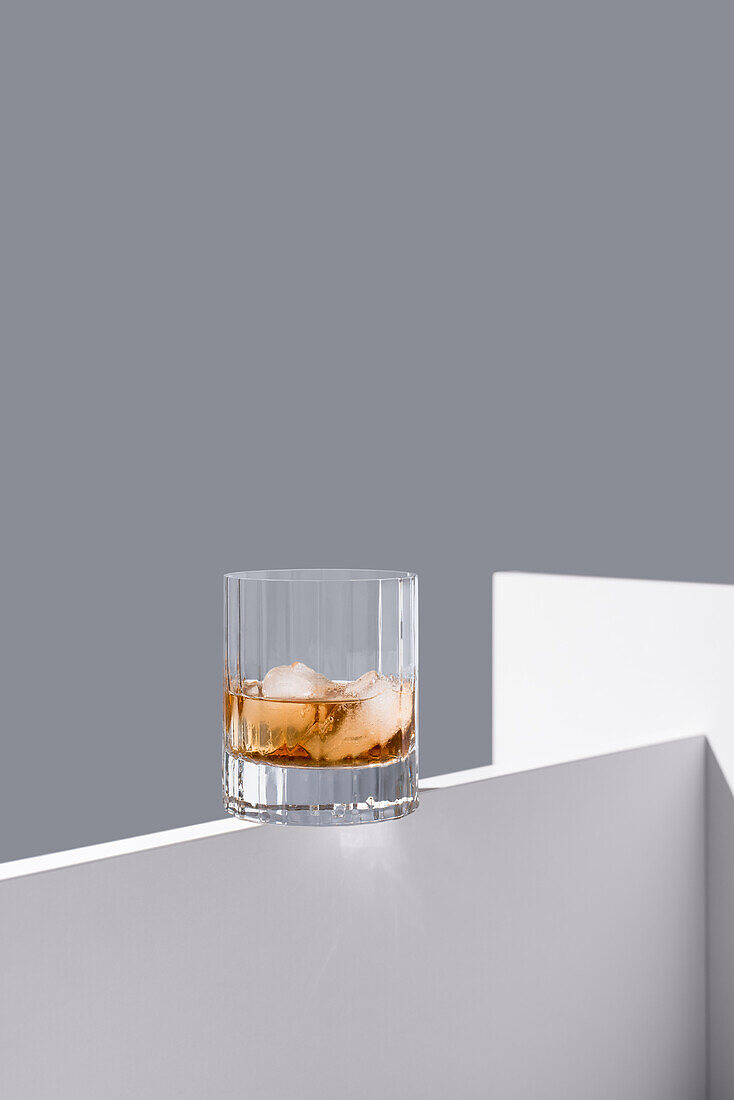 Transparent glass of whiskey on rocks with ice cubes on a white wooden table isolated on a grey background