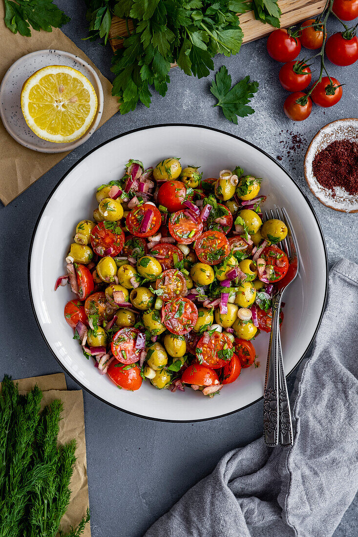 Green olive salad in a white bowl, forks inside, sumac, cherry tomatoes, parsley and dill on the side