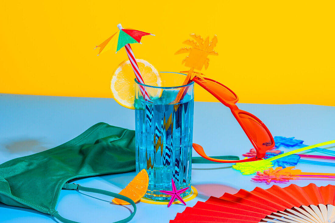 Composition of cocktail glass and sliced orange fruit with straw, red glasses on surface with inner wear, hand fan on blue and yellow background