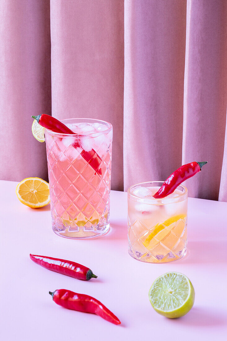 Transparent glasses filled with cold, refreshing cocktails, served with chilli peppers and ice cubes, on a table with lemons and limes