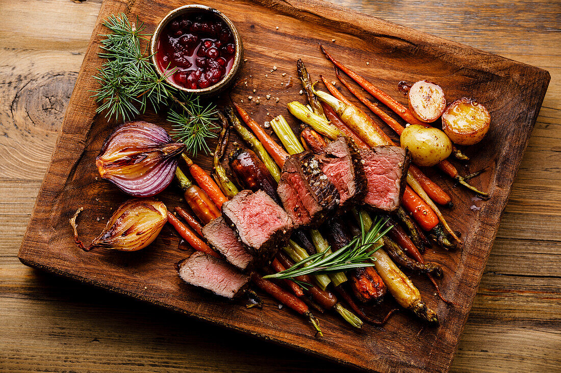 Grilled venison steak with baked vegetables and berry sauce on a wooden base