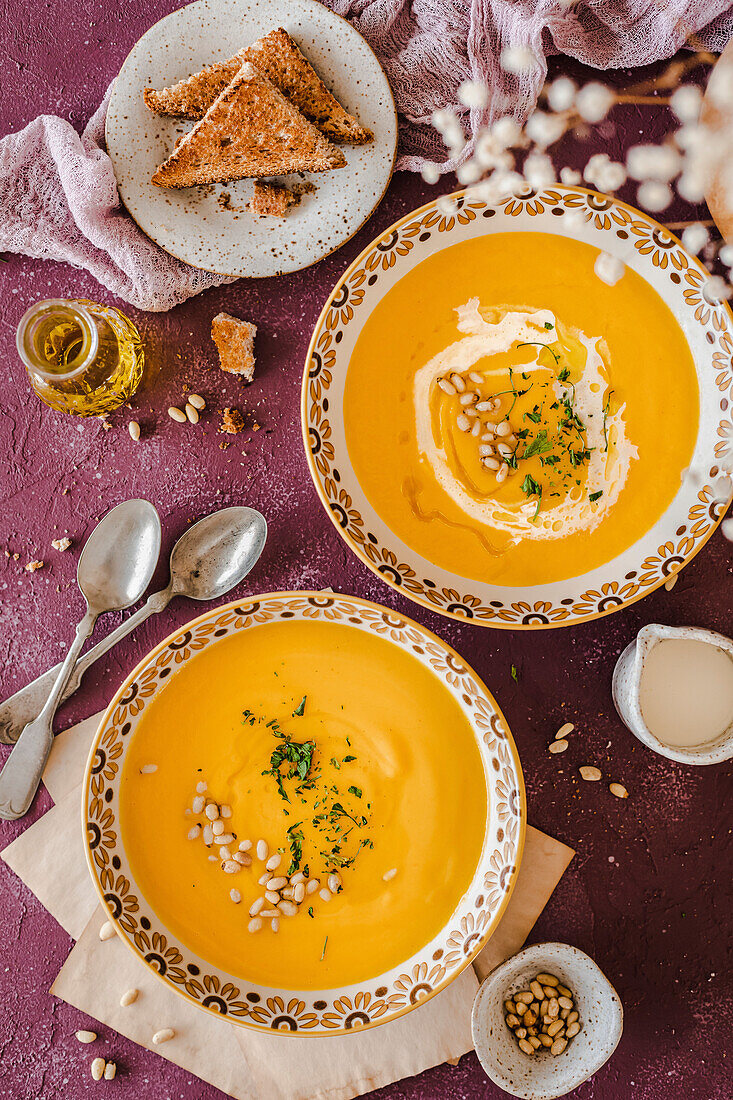 Two bowls of pumpkin soup served with brown toast and garnished with cream, pine nuts and herbs on a purple background