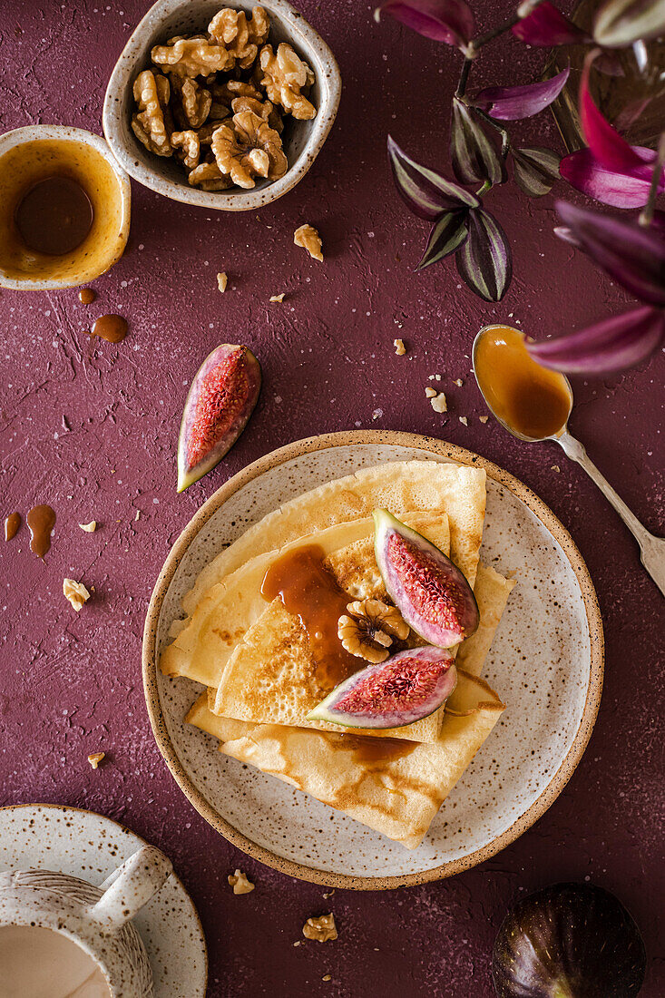 Crepes folded on a handmade ceramic plate, decorated with figs, caramel and walnuts on a purple background