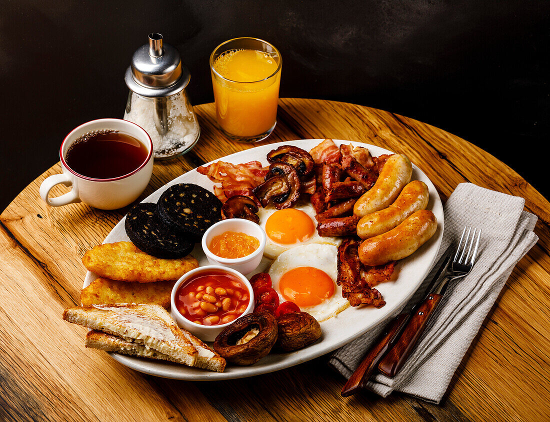 A full English breakfast with fried eggs, sausages, bacon, black pudding, beans, toast and tea on a wooden base
